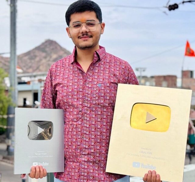 Tarjan Khatri smiling outdoors in Rajasthan, holding a YouTube Silver Play Button in one hand and a YouTube Gold Play Button in the other, with a hill in the background
