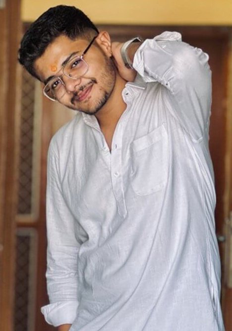 Tarjan Khatri in a white kurta and glasses, posing by a wooden door with one hand behind his head